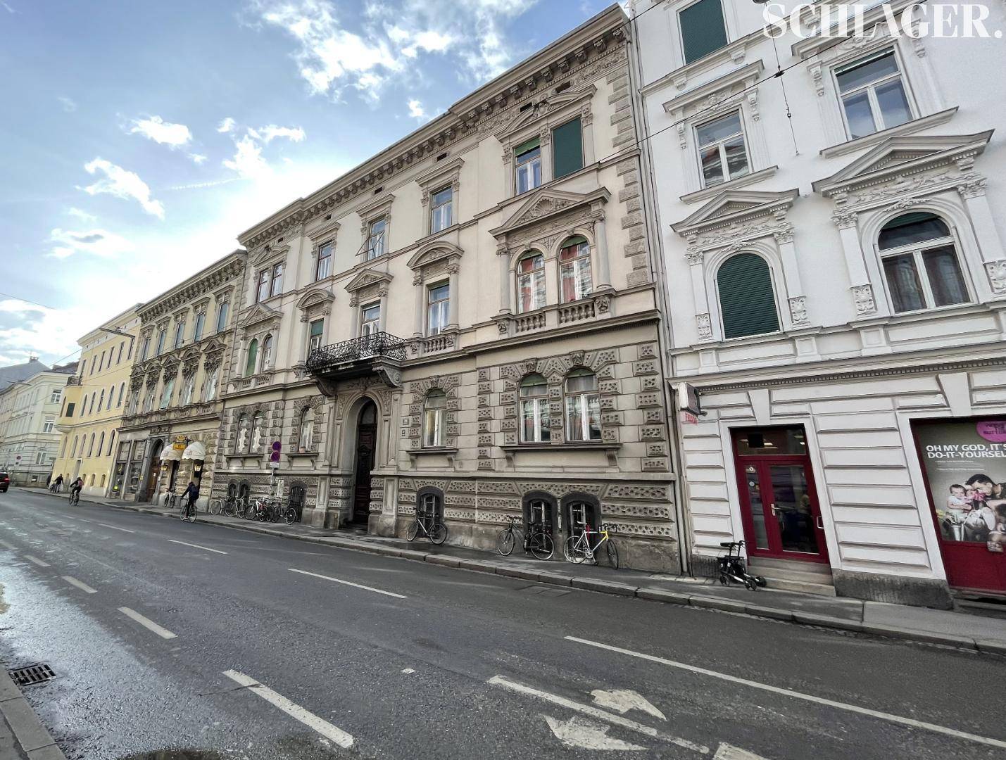 Breite Ansicht eines historischen Stadtgebäudes mit klassischer Architektur und angrenzenden Häusern an einer Straße.