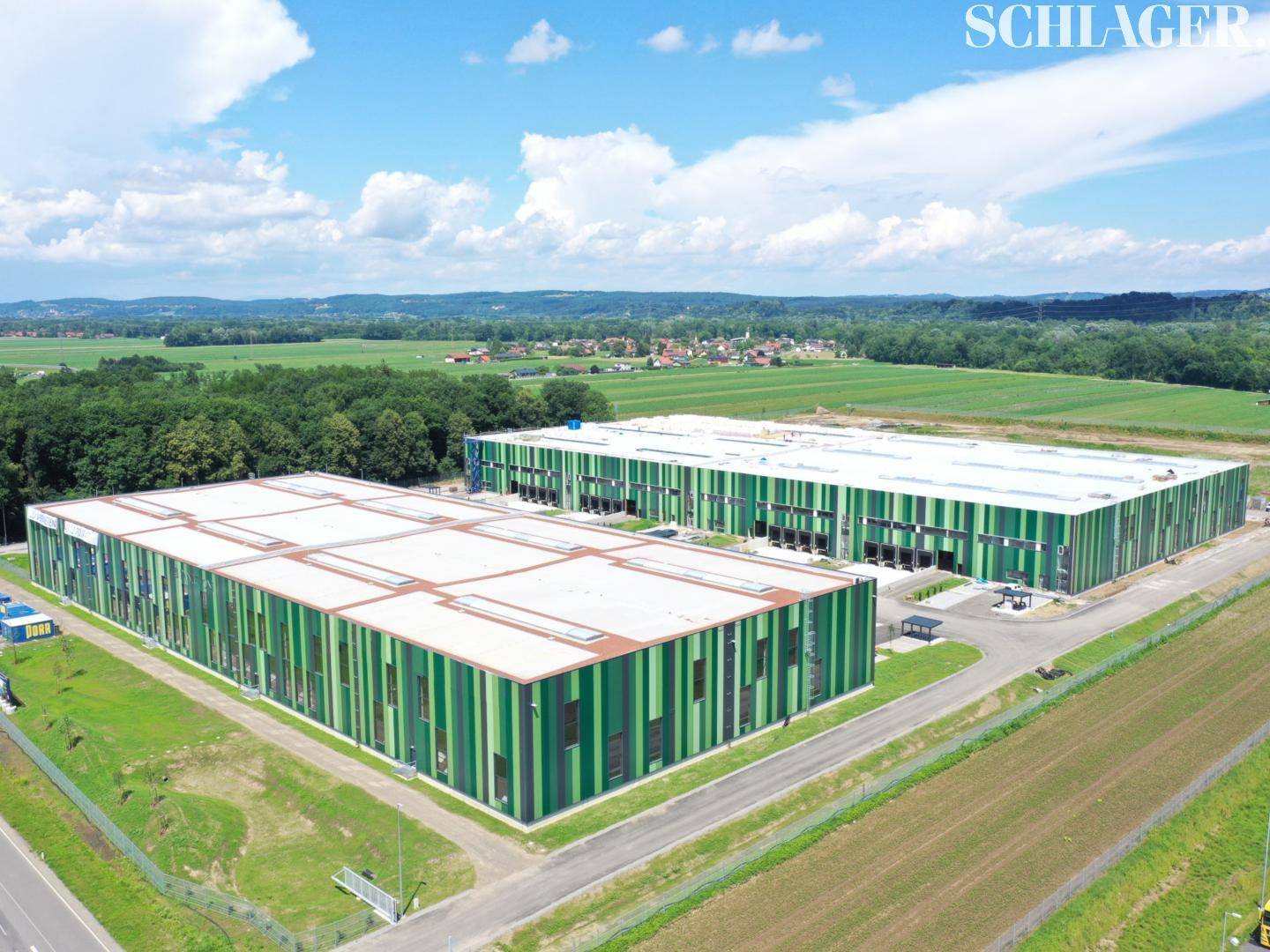 Luftaufnahme von zwei großen, modernen Industriegebäuden in einer ländlichen Umgebung mit viel Grün und Feldern.