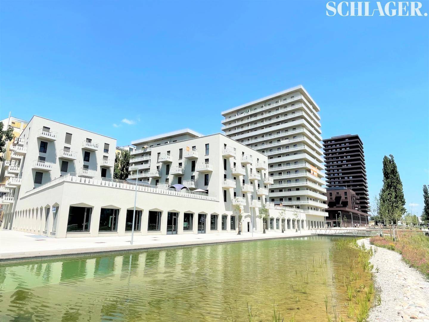 Moderne Wohngebäude entlang eines Wasserlaufs mit Grünflächen und Bäumen unter strahlend blauem Himmel.