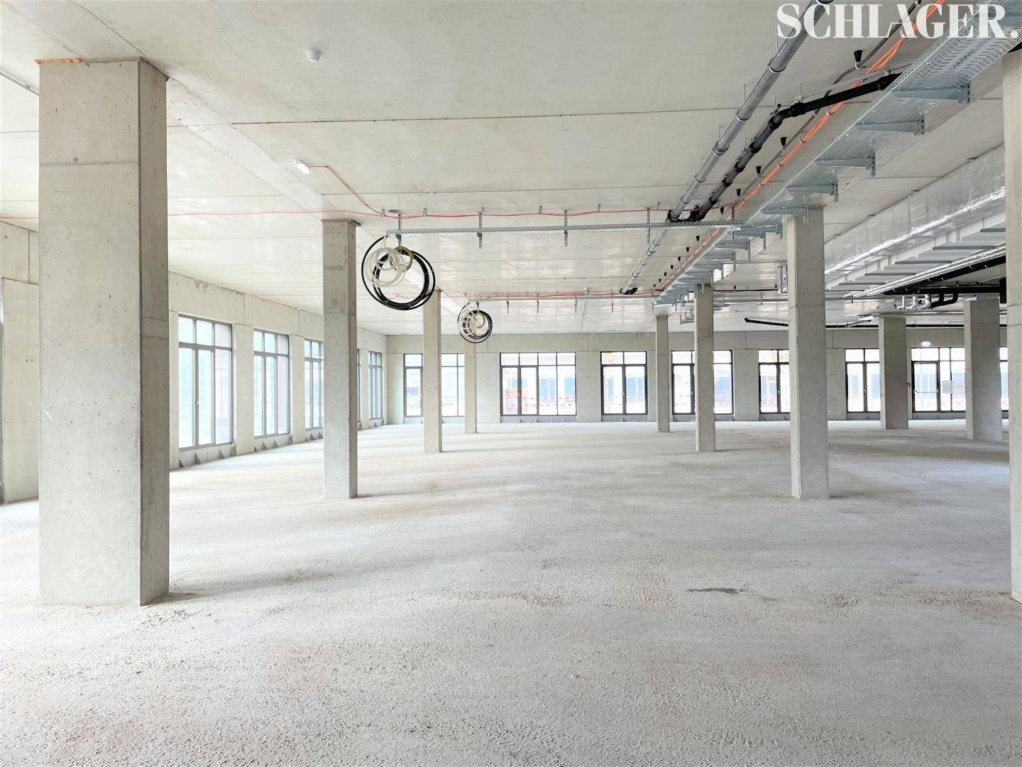 Großer, offener Gewerberaum im Rohbau mit vielen Fenstern, Betonsäulen und sichtbaren Installationen an der Decke.