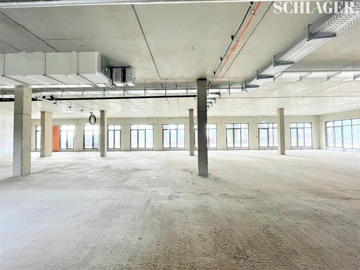 Heller, weitläufiger Innenraum im Rohbau mit vielen Fenstern und sichtbaren Deckeninstallationen, bereit für den Ausbau.