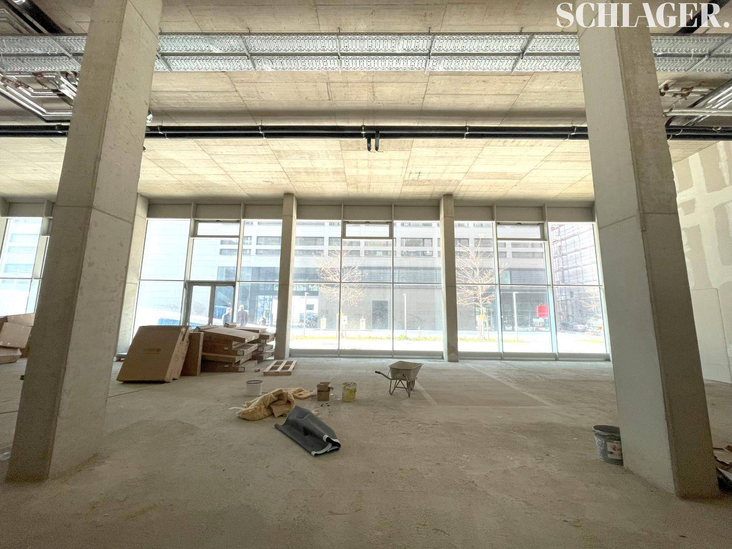 Geräumiger, unfertiger Gewerberaum mit hohen Decken, Betonpfeilern und großen Fensterfronten, viel Tageslicht.