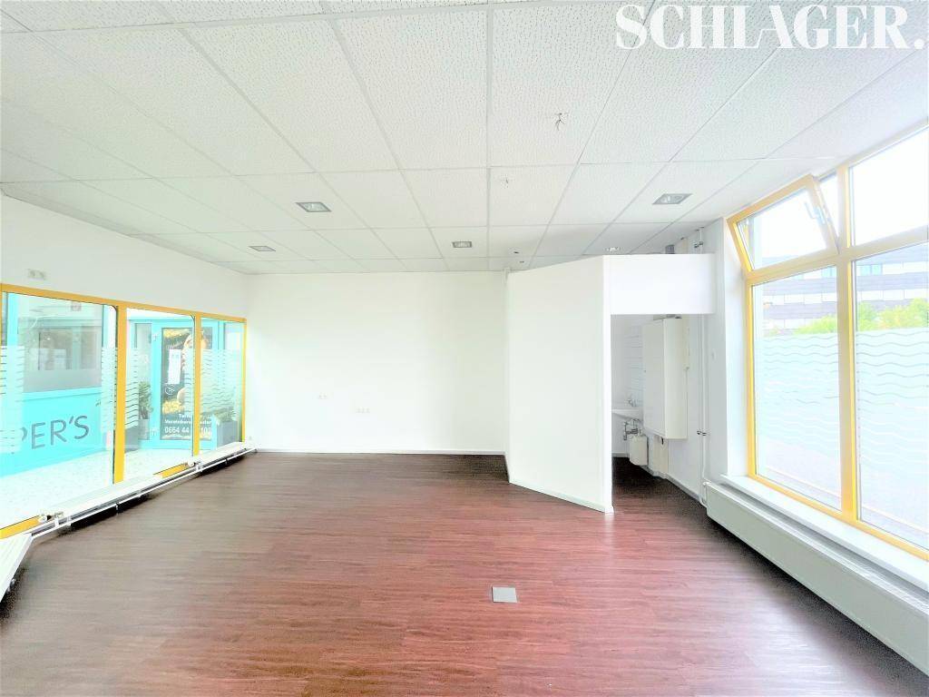 Heller Gewerberaum mit großen Fenstern, dunklem Boden und einem kleinen Sanitärbereich im Hintergrund.