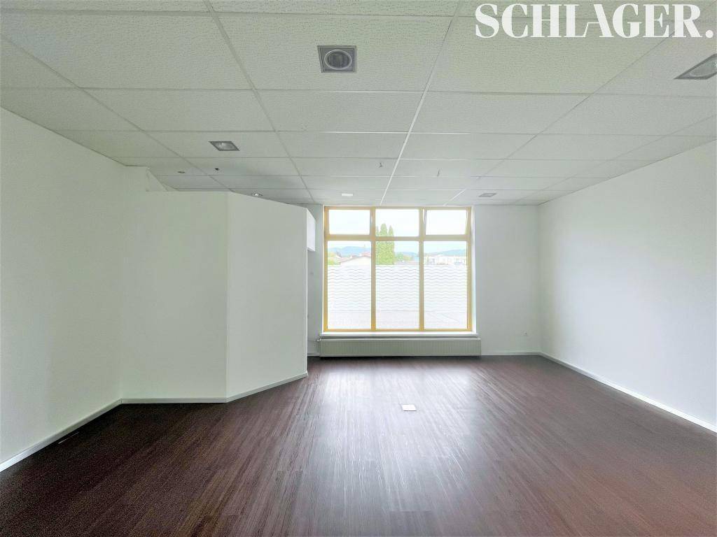 Leerer Gewerberaum mit dunklem Laminatboden, weißen Wänden und einem Fenster mit Blick ins Grüne.