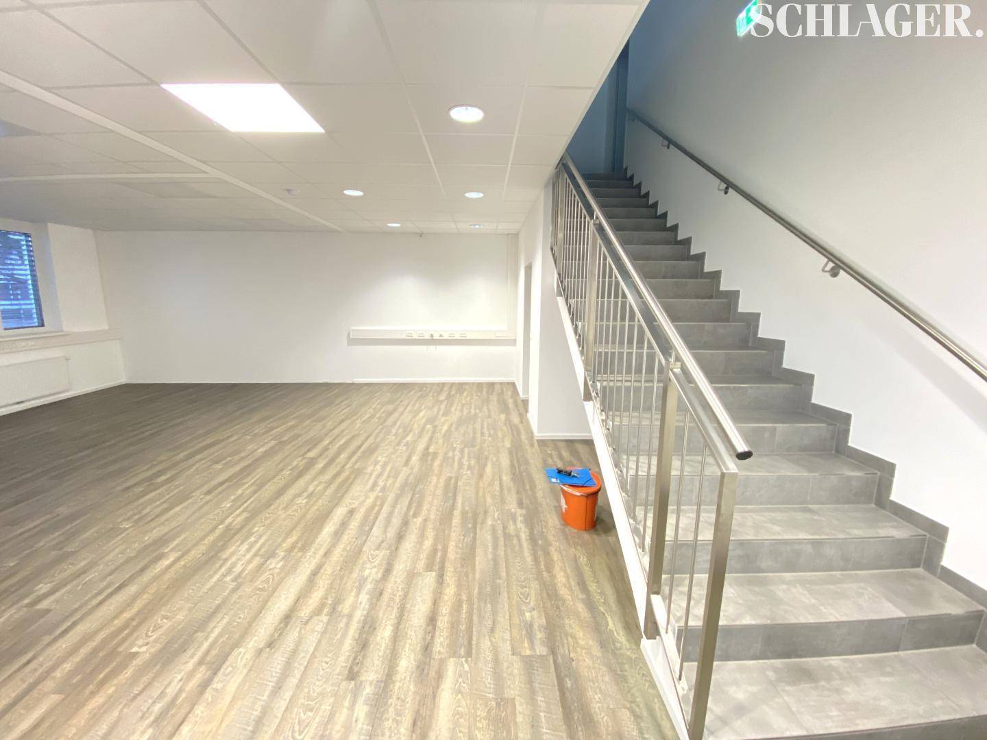 Geräumiger Bürobereich mit hellem Laminatboden und einer angrenzenden Treppe mit Metallgeländer.