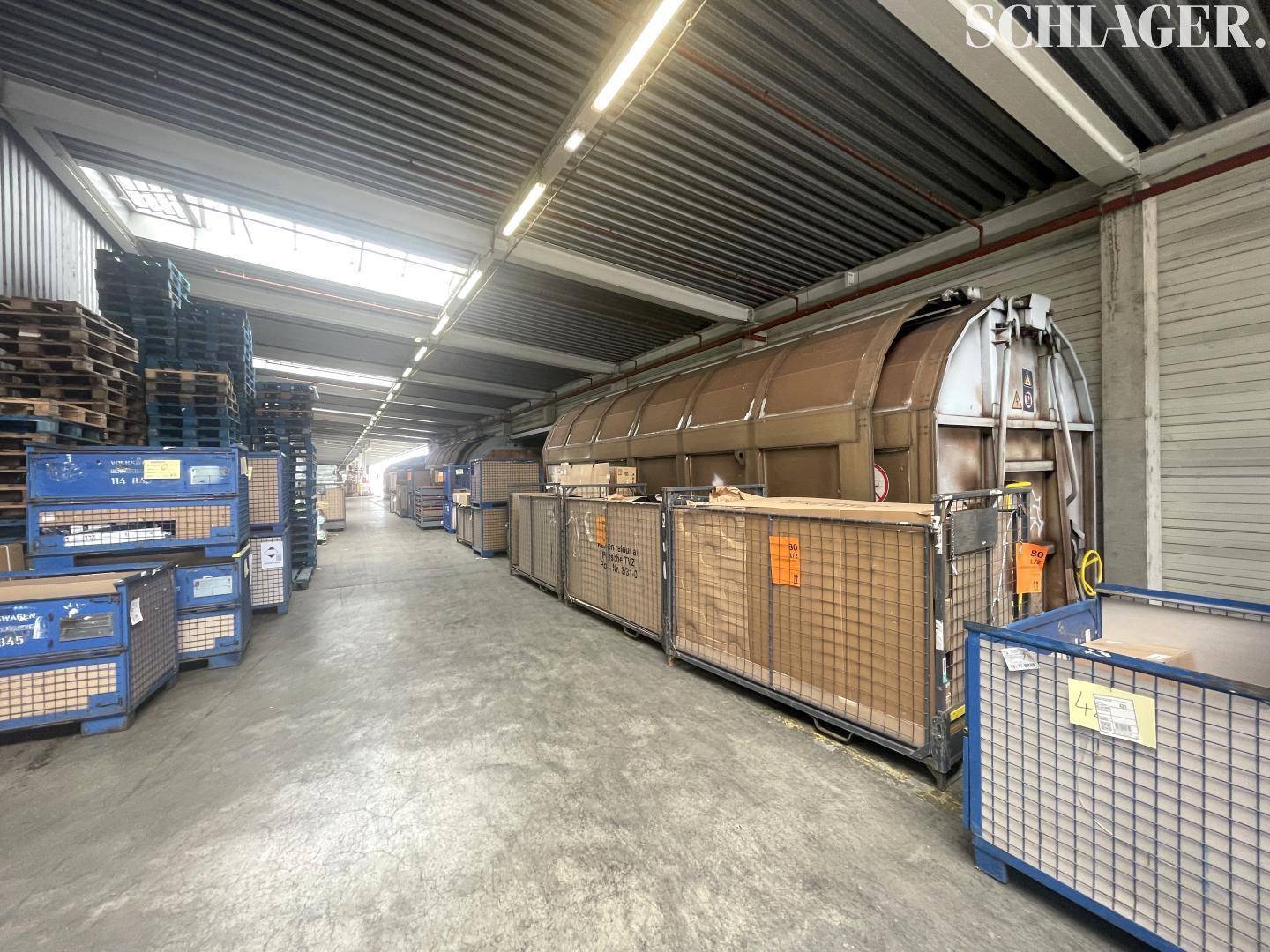 Langer Gang in einer Lagerhalle mit Rollcontainern, Paletten und Oberlichtern für Tageslicht.