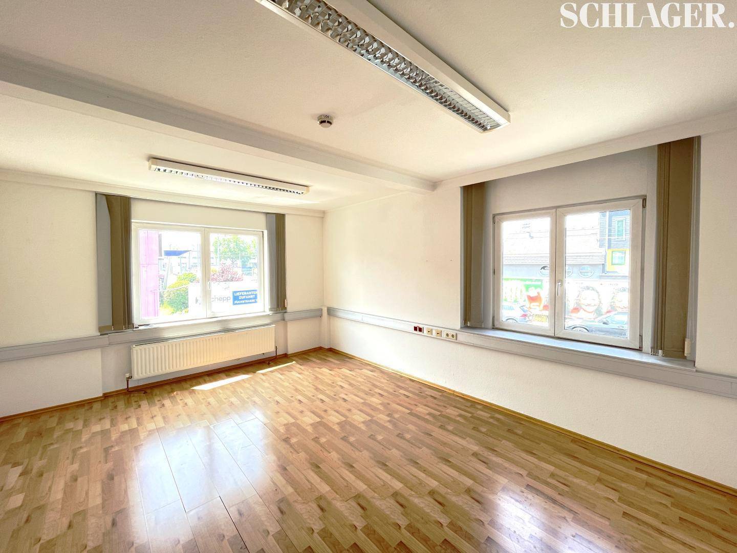 Geräumiger Büroraum mit zwei großen Fenstern, Laminatboden und weißen Wänden.