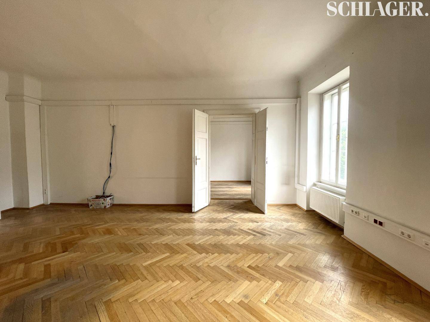 Heller, leerer Raum mit Fischgrätparkettboden, einem Fenster und Zugang zu weiteren Zimmern.