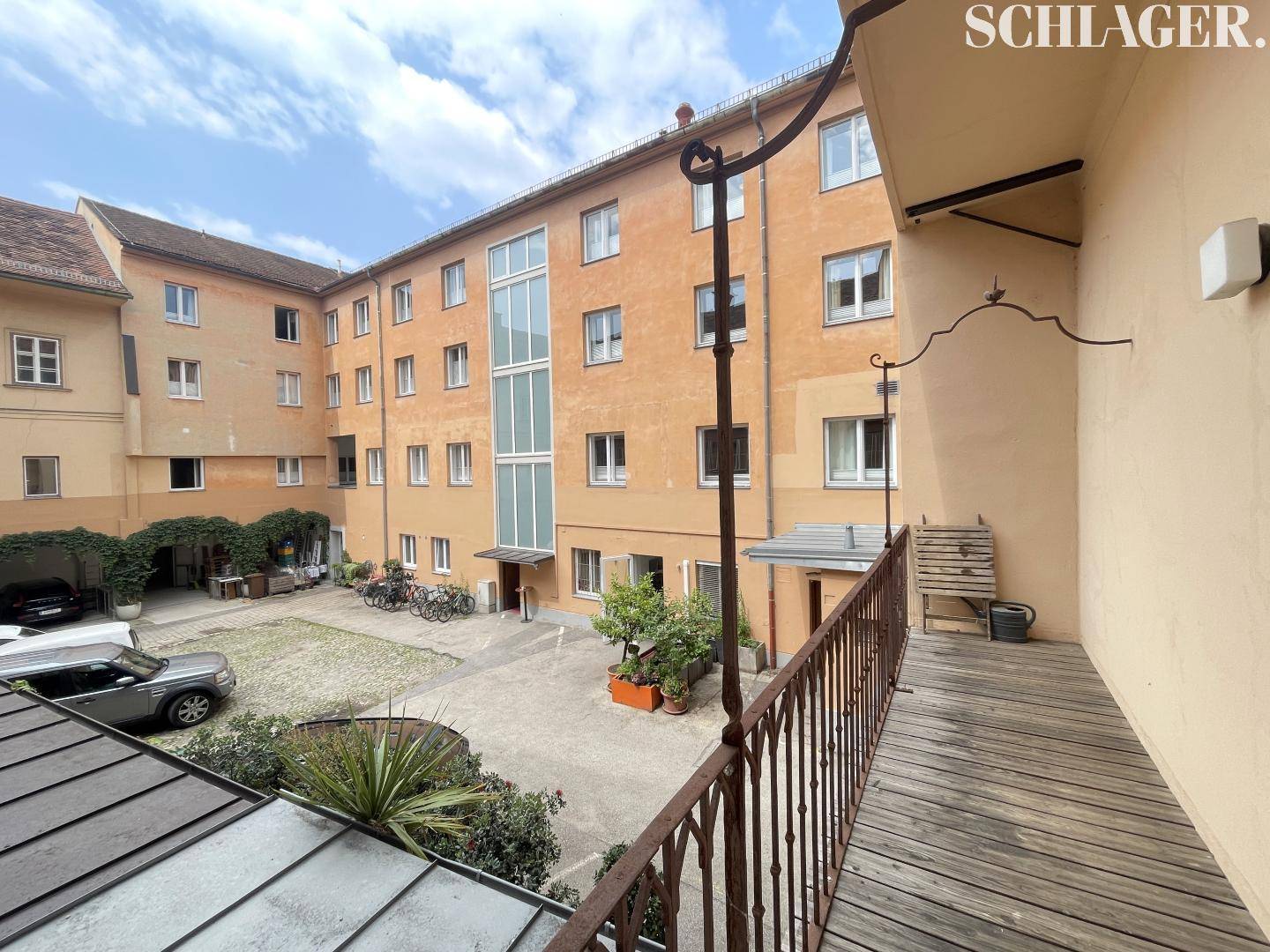 Blick von einem Balkon auf einen begrünten Innenhof mit parkenden Autos und Fahrrädern.