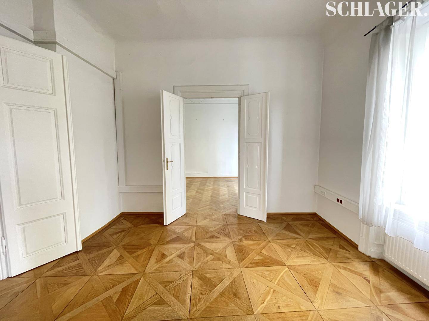 Eleganter Raum mit geometrischem Parkettboden, weißen Wänden und hohen Doppeltüren.