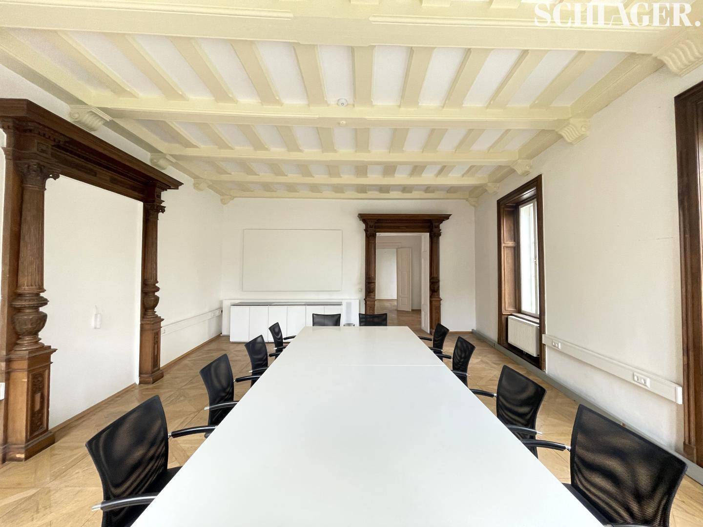 Konferenzraum mit langem Tisch, schwarzen Stühlen, Holztürrahmen und einer Kassettendecke.
