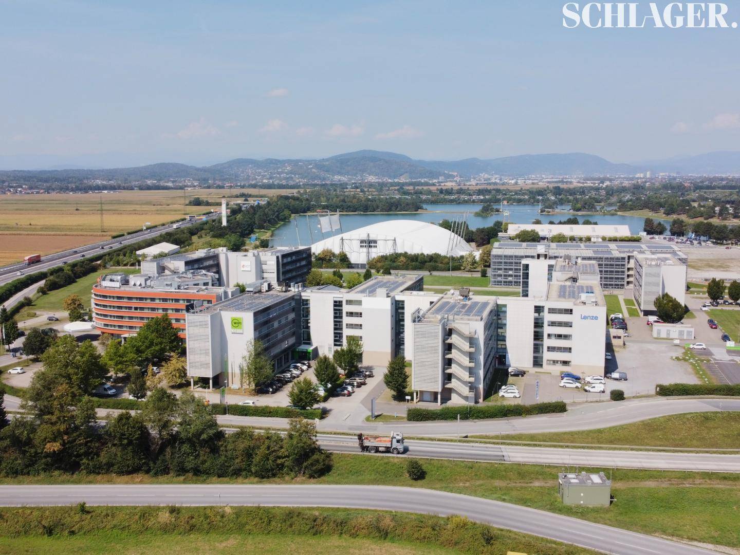 Luftaufnahme eines modernen Geschäftsparks mit See, Autobahn und Bergen im Hintergrund.