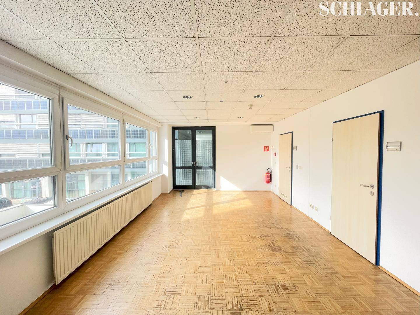 Geräumiger, leerer Büroraum mit Fischgrätparkett, großen Fenstern und abgehängter Decke.