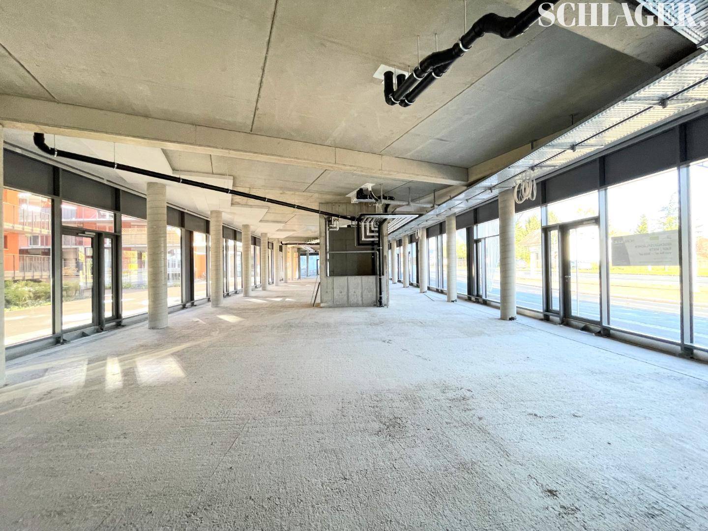 Weitläufiger, unfertiger Gewerberaum mit Betonoberflächen, freiliegenden Leitungen und viel Tageslicht durch große Fenster.