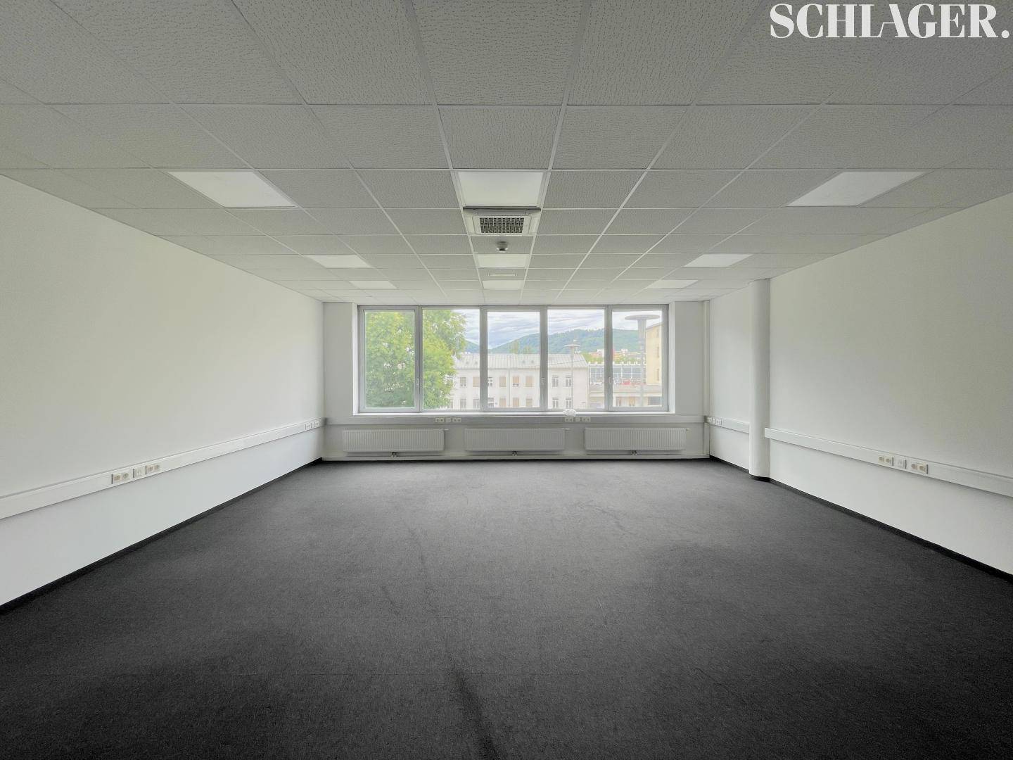 Geräumiges, leeres Büro mit Teppichboden, großen Fenstern und einem Ausblick auf grüne Hügel.