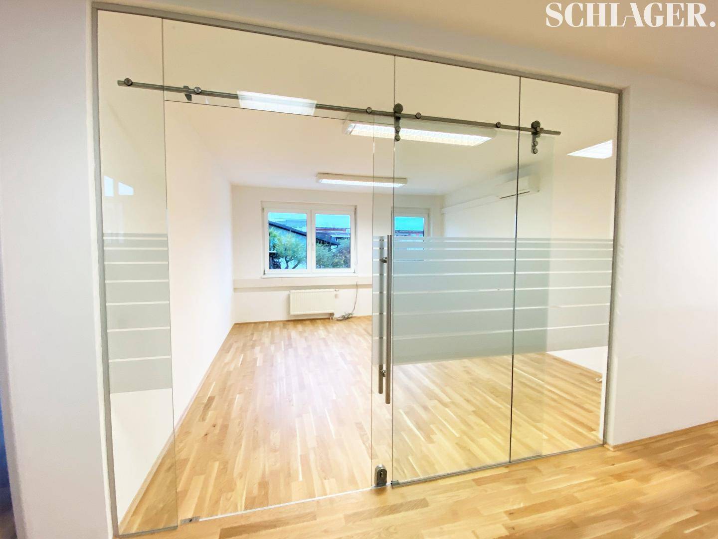 Blick durch eine moderne Glasschiebetür mit Milchglasstreifen in einen hellen Büroraum mit Holzfußboden.