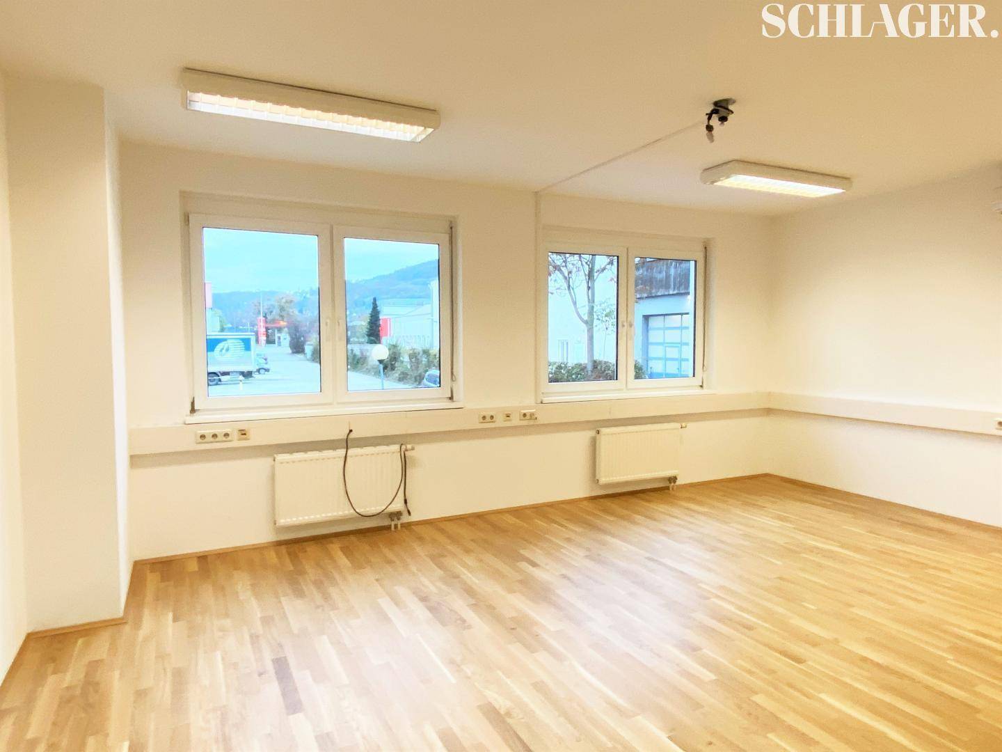 Geräumiger, leerer Büroraum mit hellem Holzfußboden, weißen Wänden und Fenstern mit Blick nach draußen.