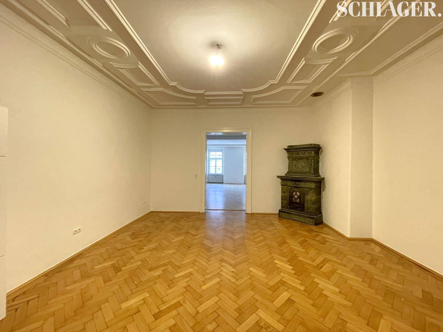 Eleganter Wohnraum mit Fischgrätparkett, Stuckdecke und einem dekorativen grünen Kachelofen.
