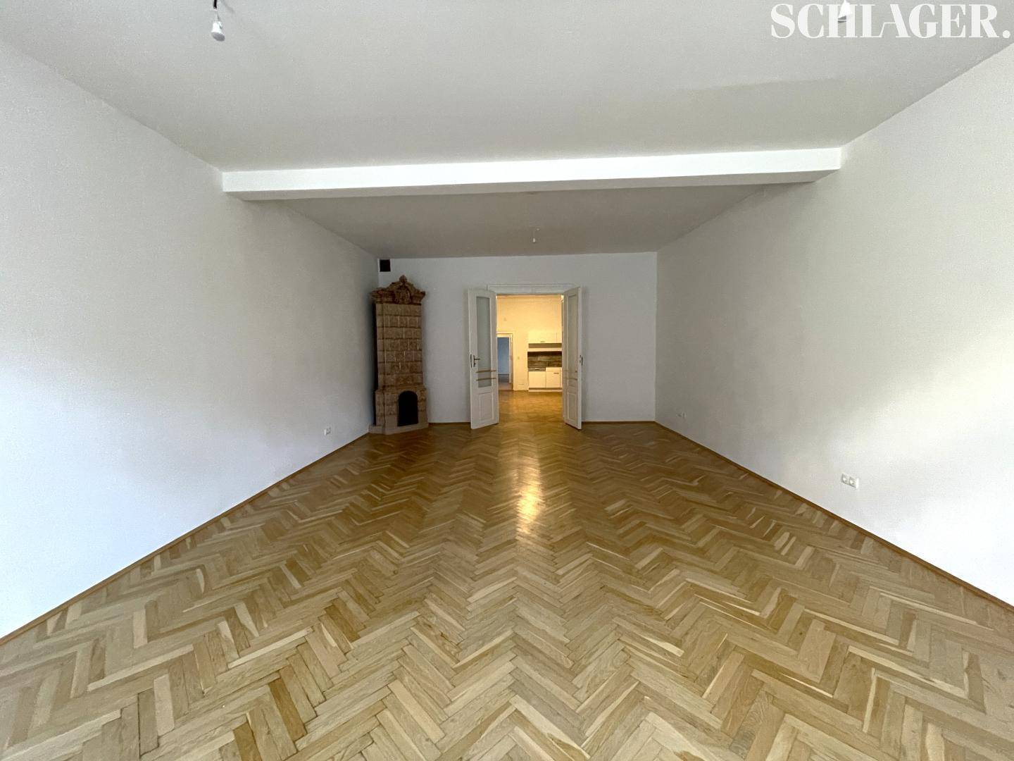 Heller Wohnraum mit traditionellem Kachelofen, Fischgrätparkett und Zugang zu einem weiteren Zimmer.