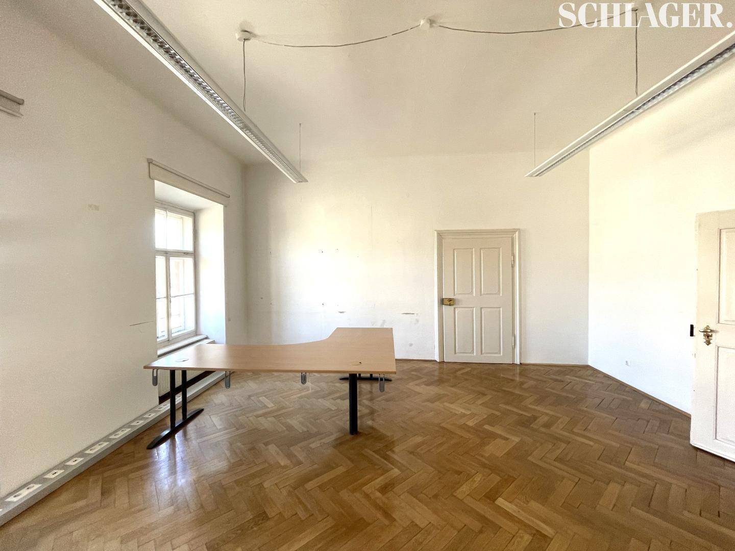 Büroraum mit Fischgrätparkett, hohen Decken und einem Schreibtisch am Fenster.