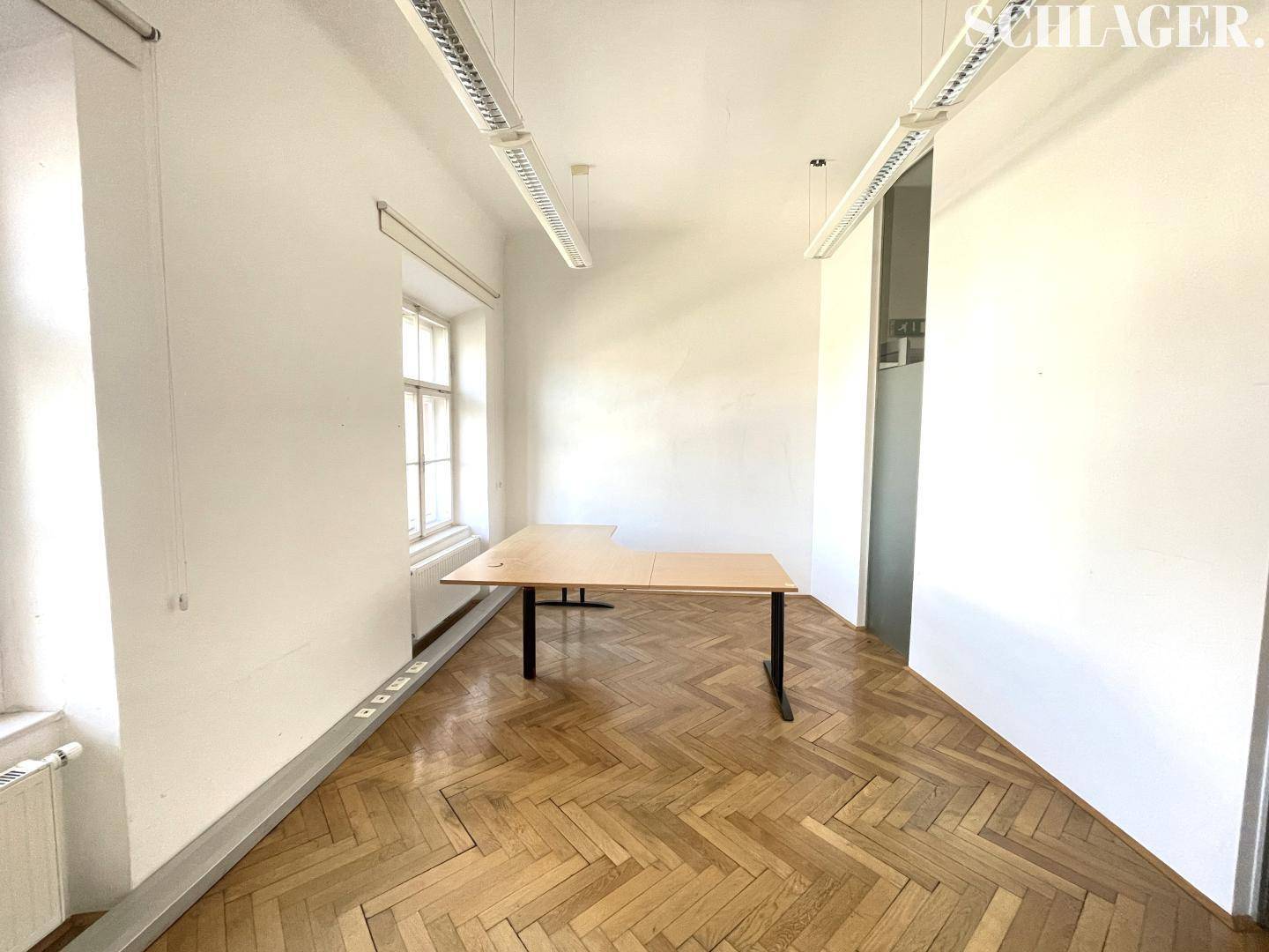 Geräumiger Büroraum mit Fischgrätparkett, hohen Decken und einem Schreibtisch.