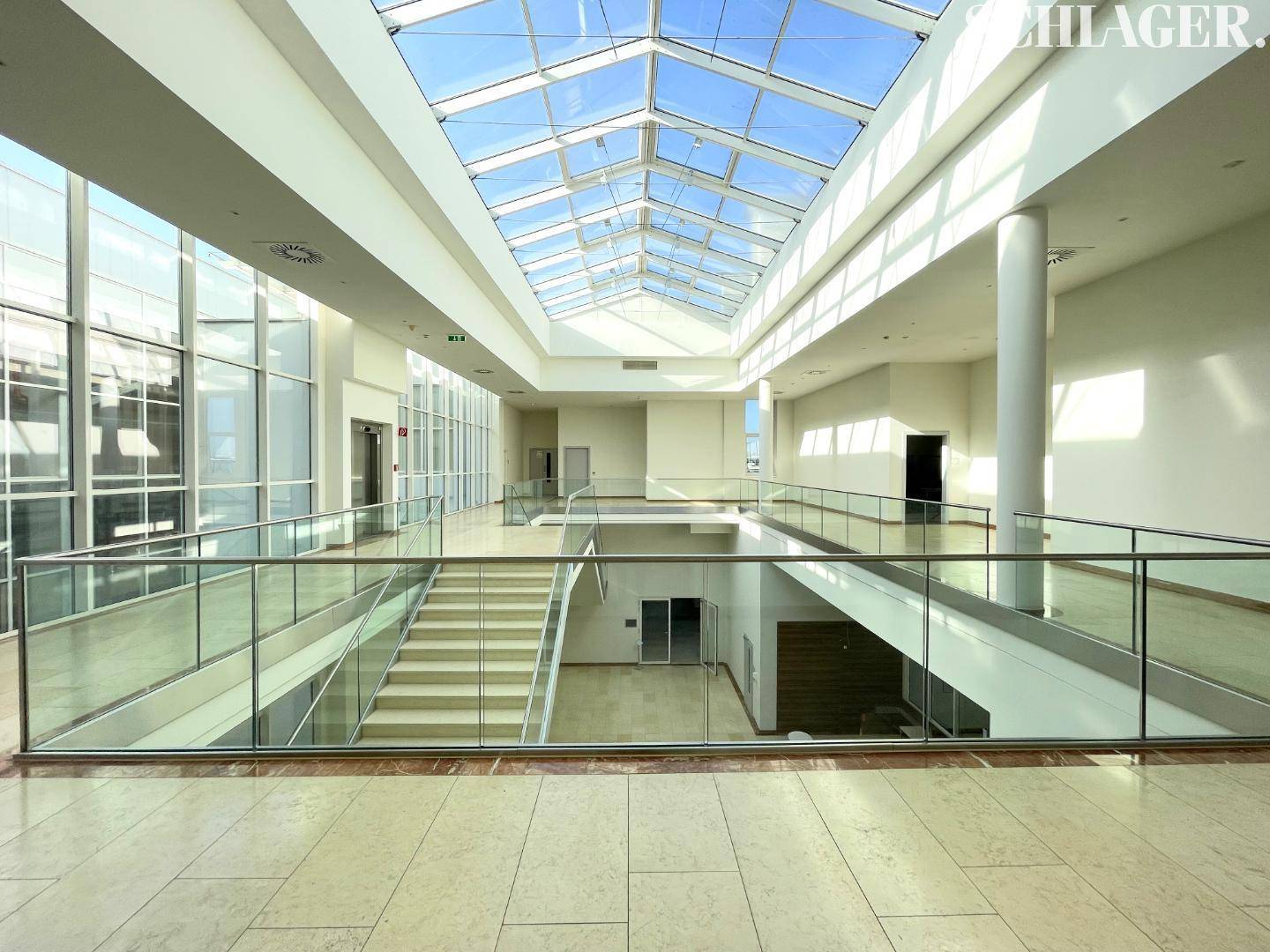 Geräumiges Atrium mit beeindruckendem Glasdach, mehreren Etagen, Treppen und modernen Glasgeländern, sehr hell.