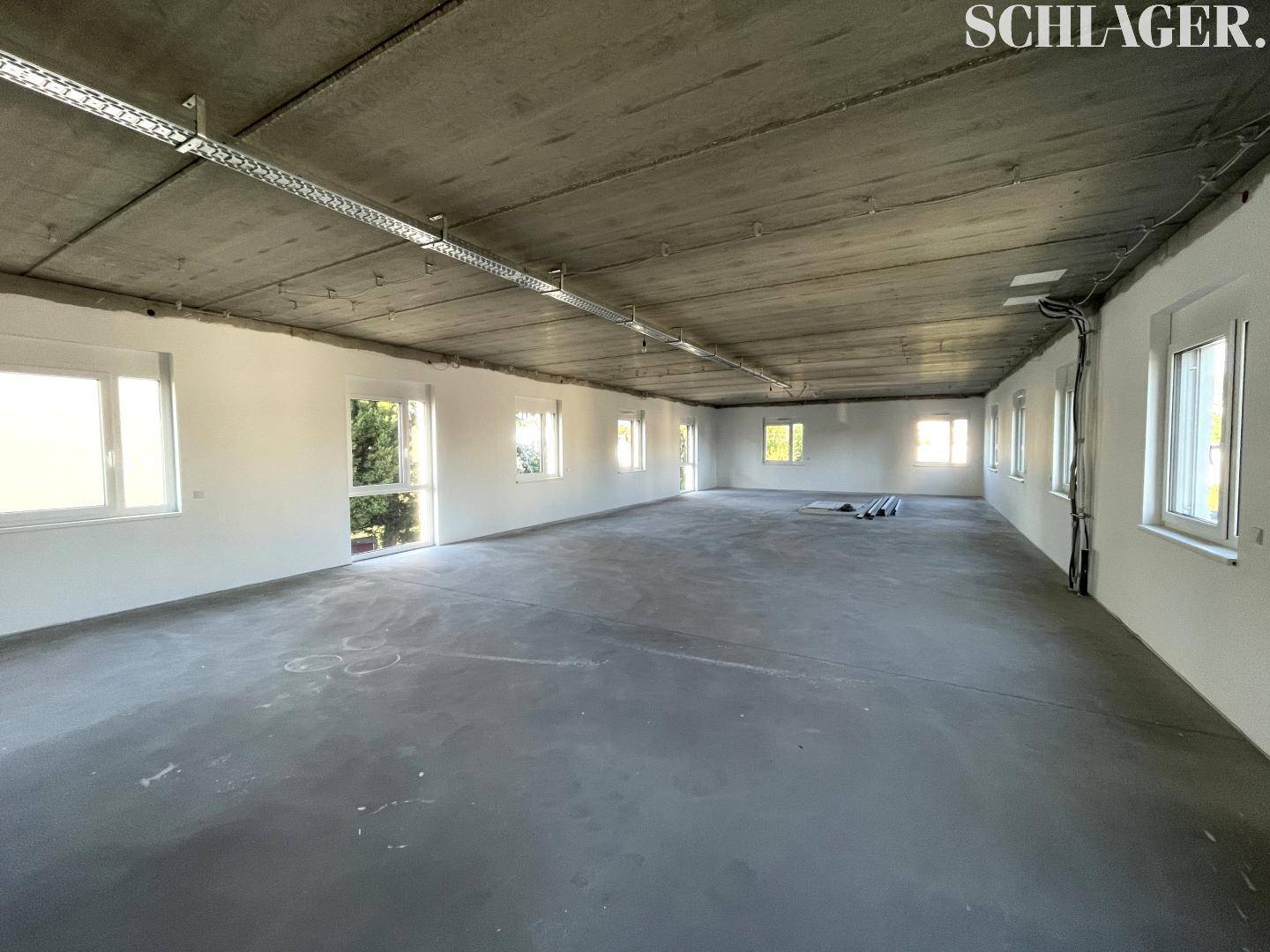 Heller, unmöblierter Raum mit Sichtbetondecke und zahlreichen Fenstern für viel Lichteinfall.