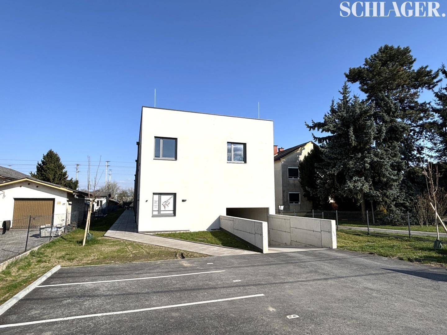 Neubau mit weißer Fassade, Tiefgaragenzufahrt und Außenparkplätzen auf einem sonnigen Grundstück.