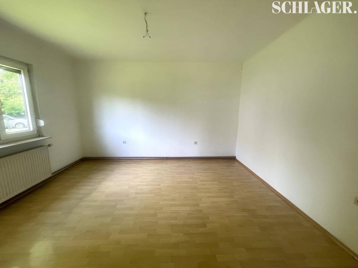 Leerer Raum mit hellem Laminatboden, weißen Wänden und einem Fenster mit Heizkörper.
