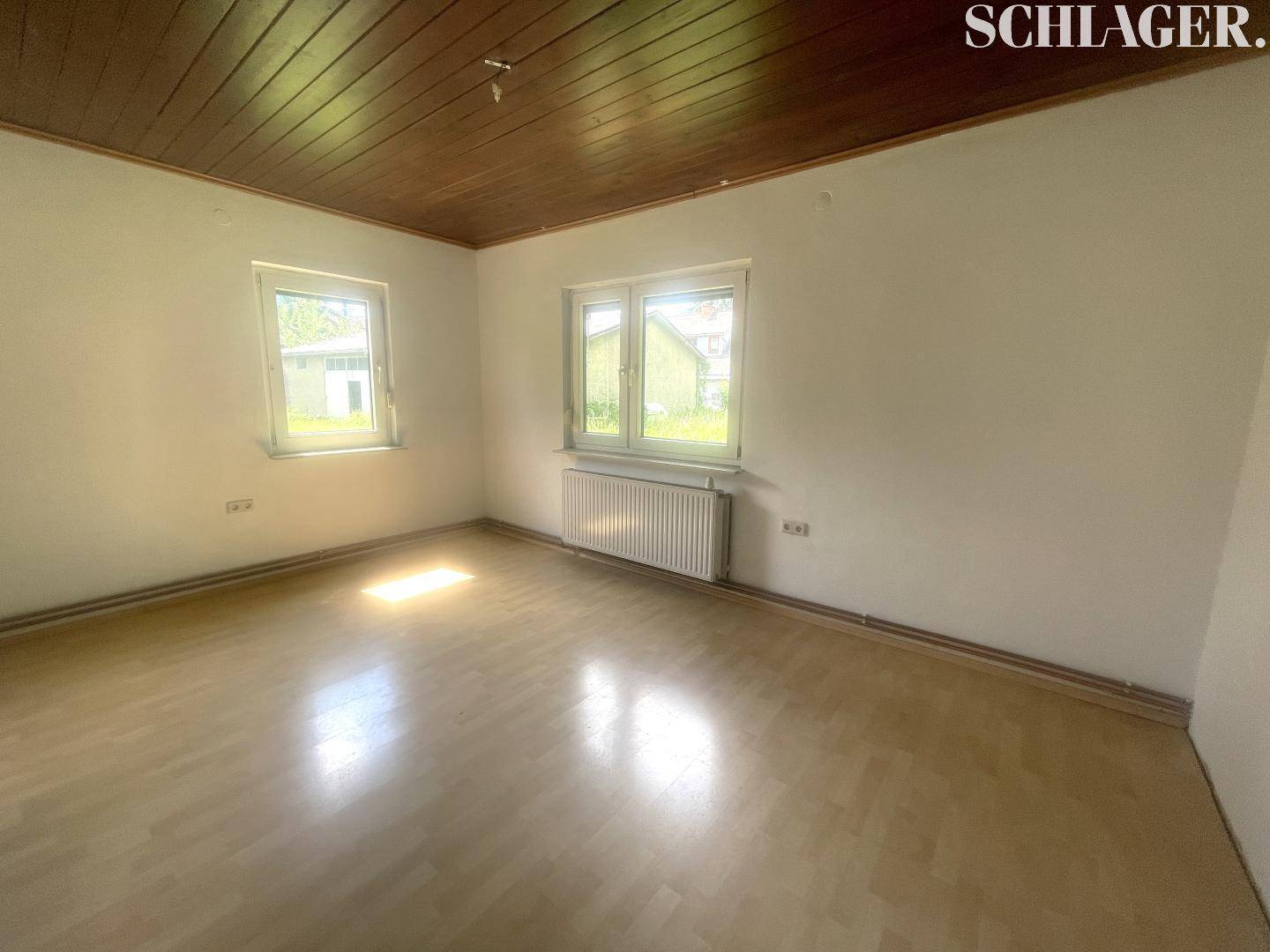 Heller, leerer Raum mit Holzdielenboden, Holzdecke und zwei Fenstern.