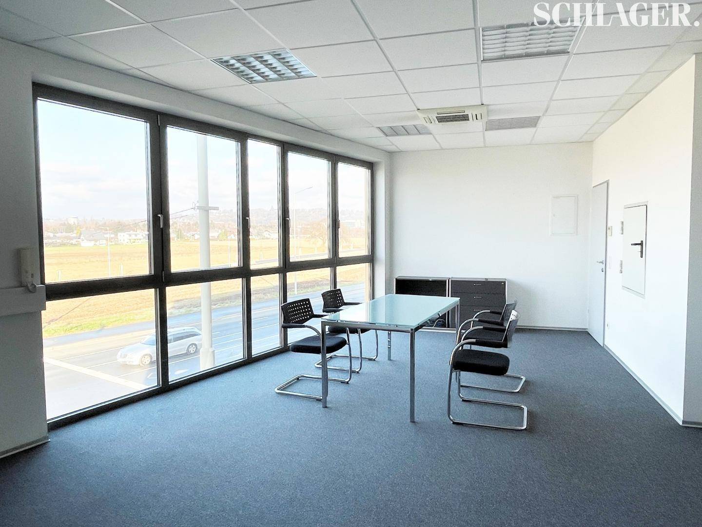 Besprechungsraum oder Büro mit Teppichboden, großen Fenstern und einem Konferenztisch mit Stühlen.
