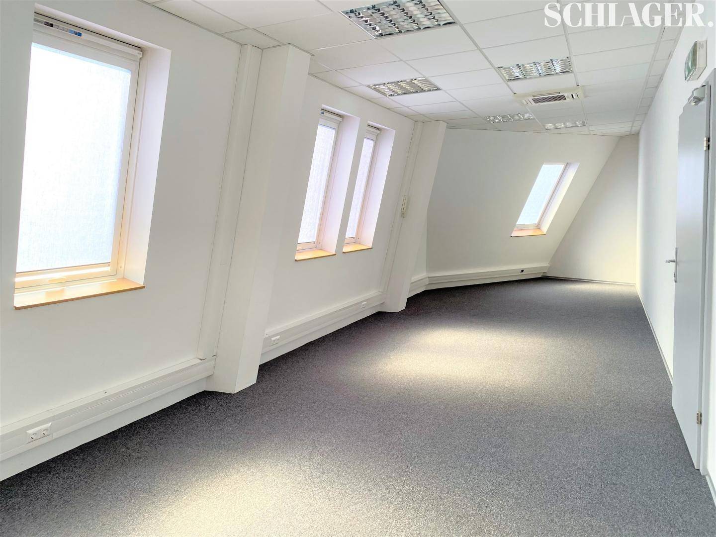 Langer Bürogang oder Arbeitsbereich mit Teppichboden, weißen Wänden und schrägen Dachfenstern für Tageslicht.