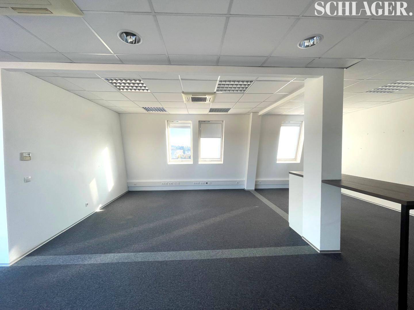 Offener Bürobereich mit Teppichboden, weißen Wänden und schrägen Dachfenstern, die viel Licht hereinlassen.