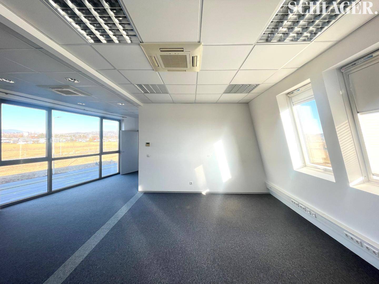 Geräumiger Bürobereich mit Teppichboden, viel Tageslicht durch große Fenster und Klimaanlage an der Decke.