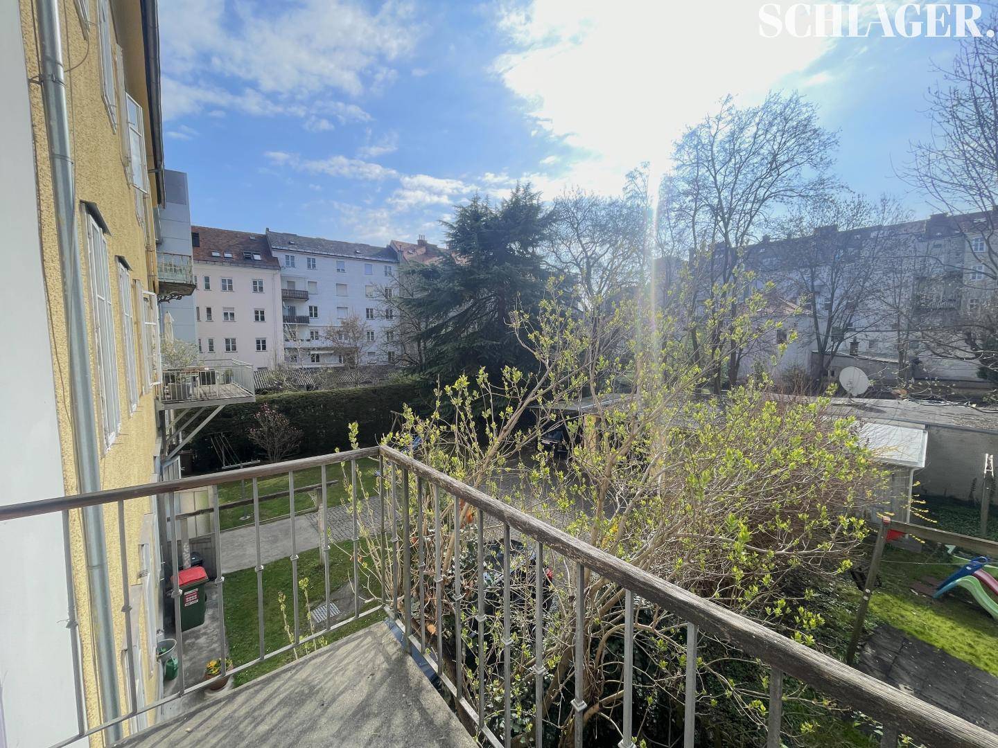 Balkon mit Metallgeländer und Blick auf einen grünen Innenhof mit Bäumen und Spielplatz.
