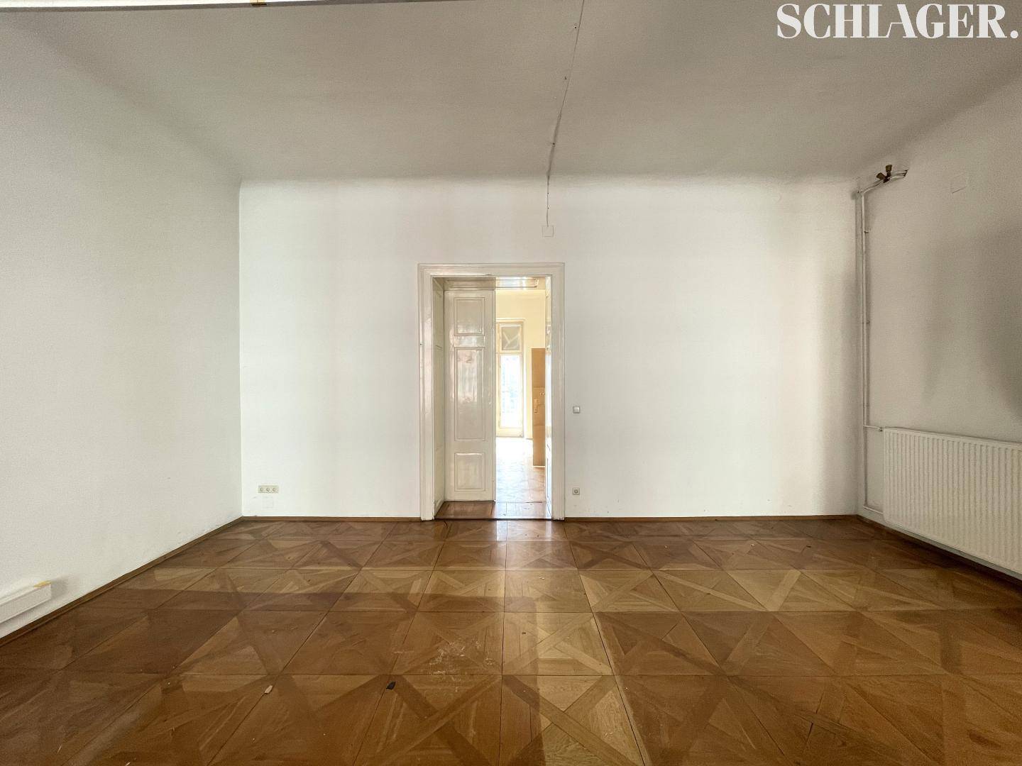 Geräumiger, leerer Raum mit klassischem Parkettboden und einer Tür zu einem weiteren Zimmer.