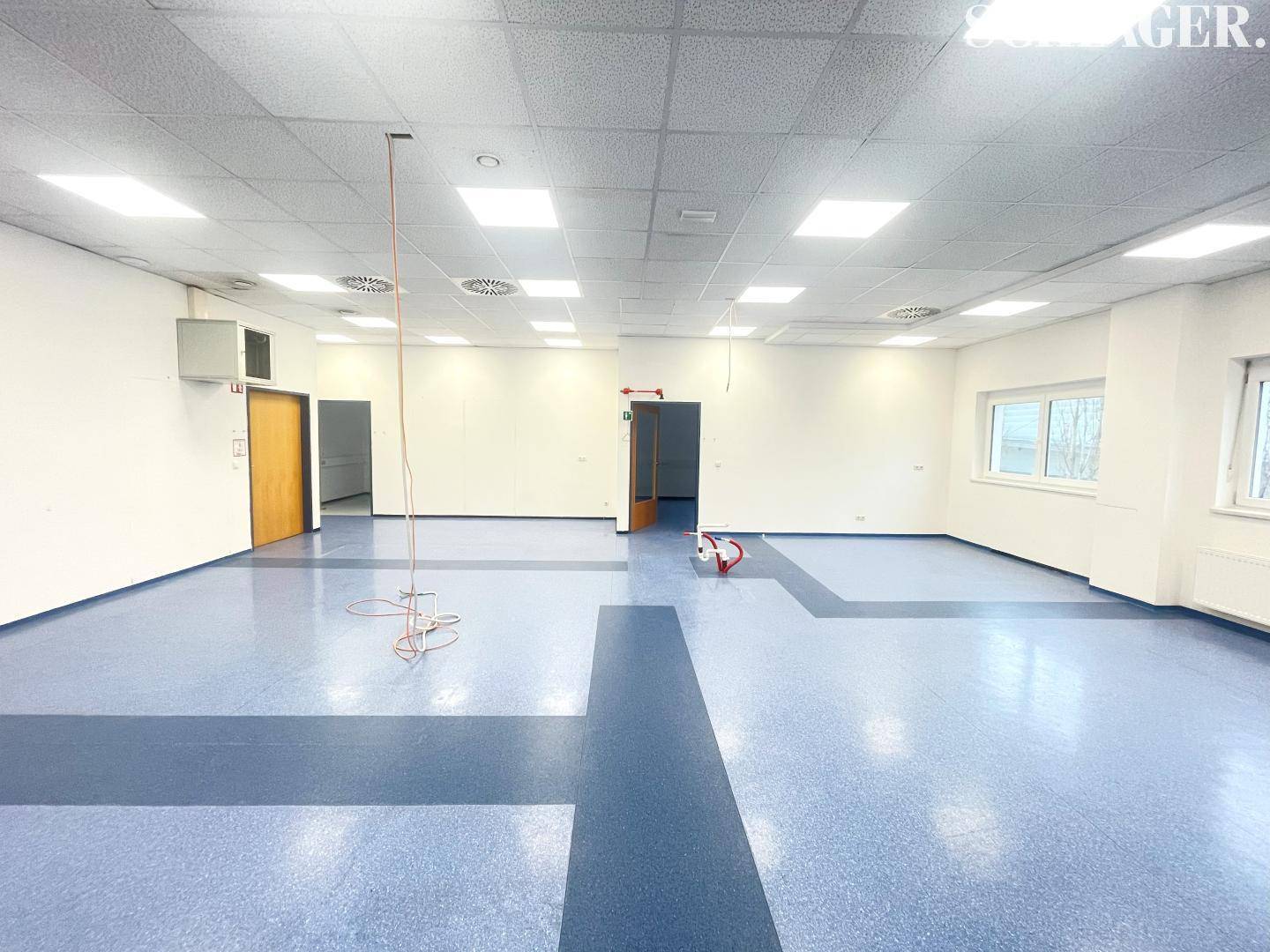 Weitläufiger Gewerberaum mit markantem blau-grauem Vinylboden, weißen Wänden und guter Beleuchtung durch Fenster.