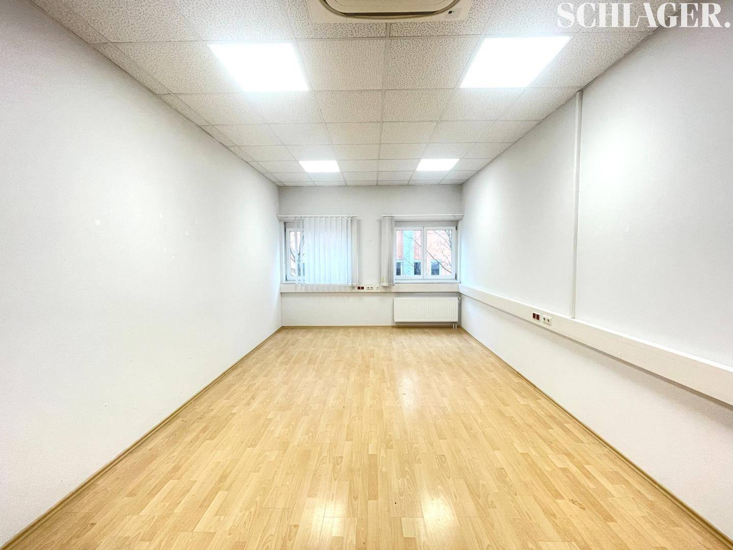 Helles, leeres Büro mit Laminatboden, weißen Wänden und Rasterdecke, mit Fenstern am Ende des Raumes.