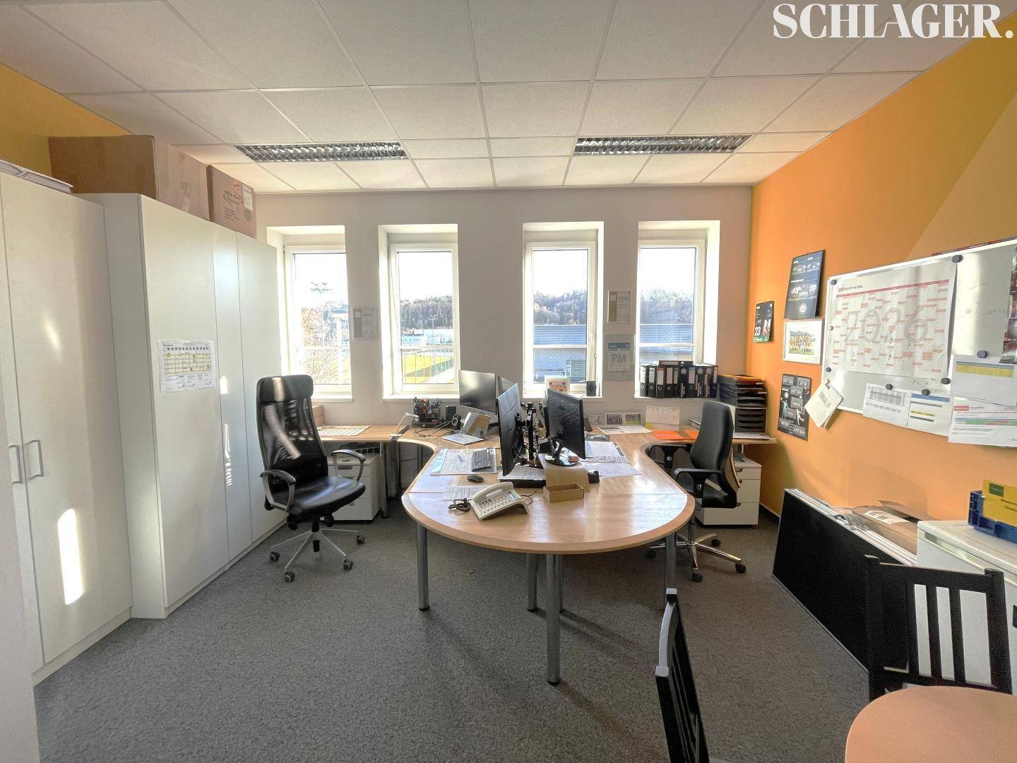 Helles Büro mit mehreren Arbeitsplätzen, weißen Schränken und Fenstern mit Blick ins Grüne.