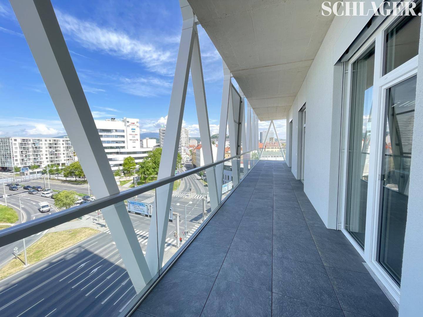 Langer, moderner Balkon mit Glasgeländer und Fliesenboden, der einen weiten Blick auf die belebte Straße bietet.