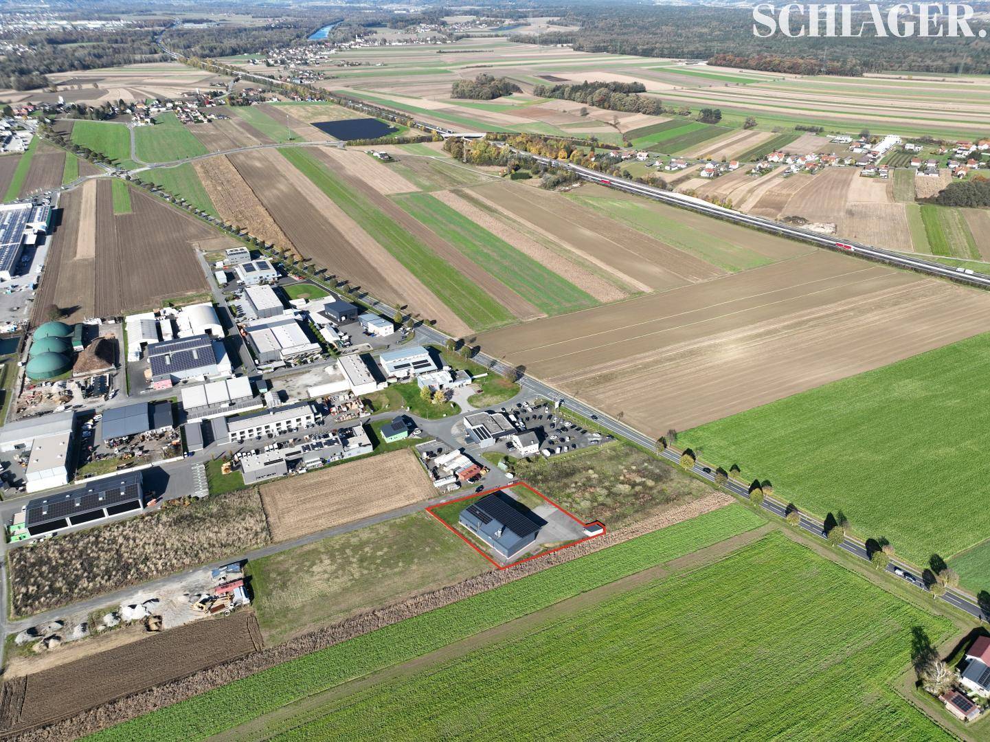 Weitläufige Luftaufnahme eines Gewerbegebiets mit Feldern, Straßen und einem Fluss in der Ferne, das Grundstück ist rot umrandet.