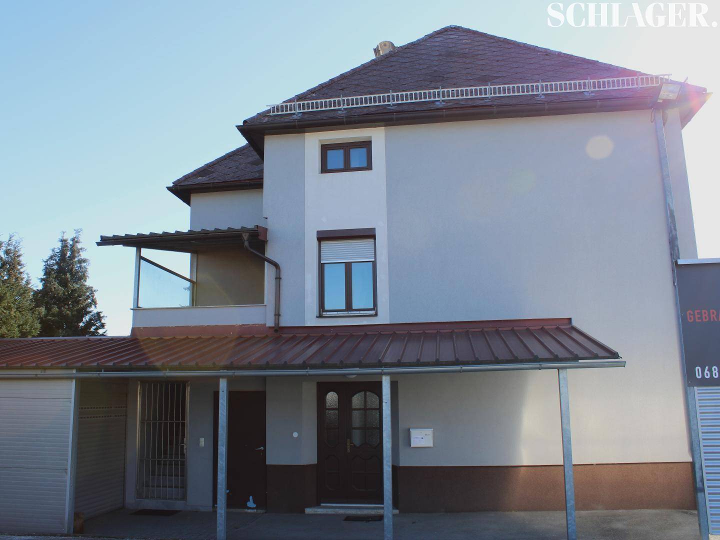 Gebäudefassade mit überdachtem Eingangsbereich und einem seitlichen, überdachten Balkon unter blauem Himmel.