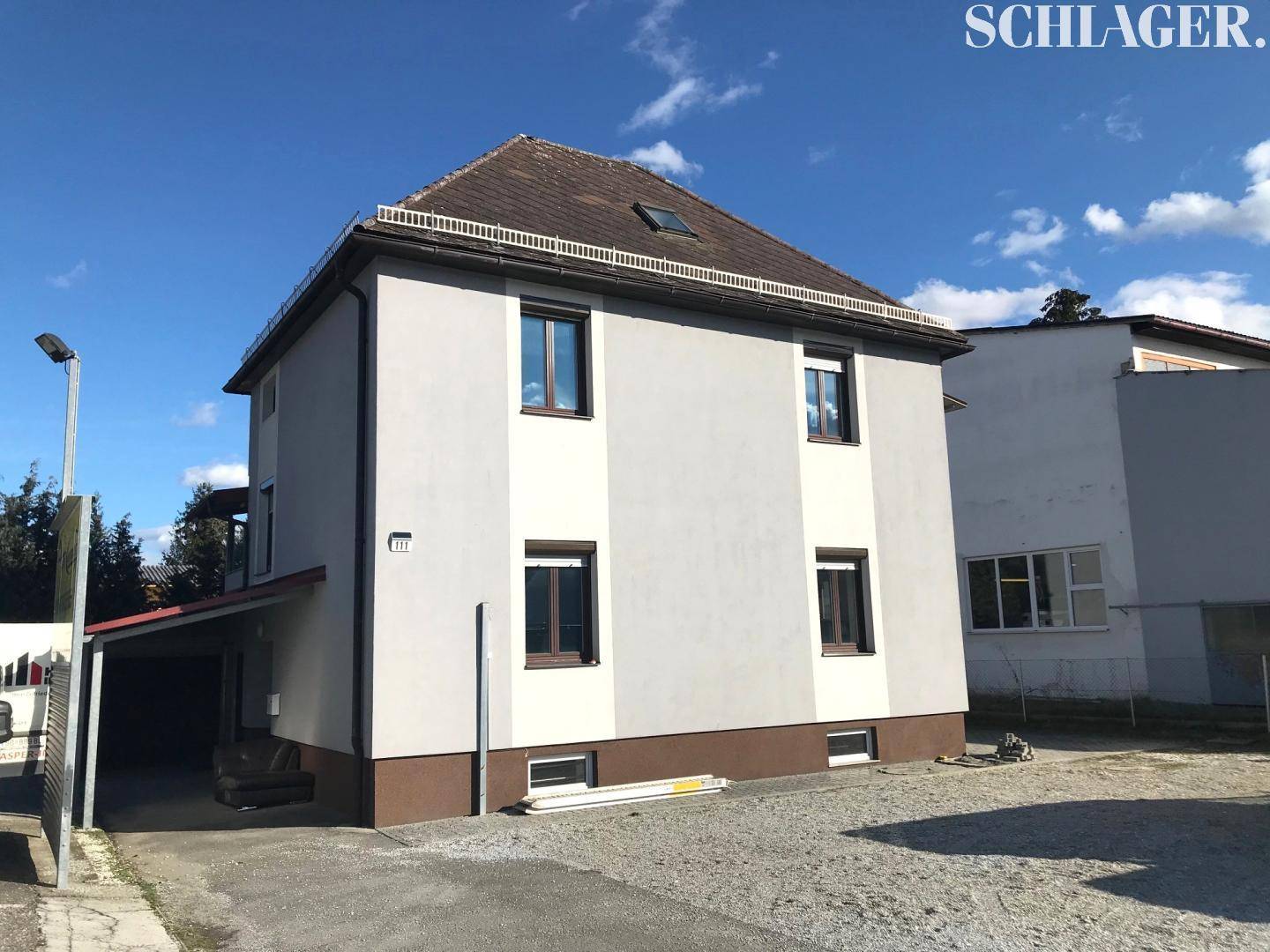 Zweistöckiges Gebäude mit hellgrauer Fassade, braunem Ziegeldach und einem Carport auf der linken Seite.