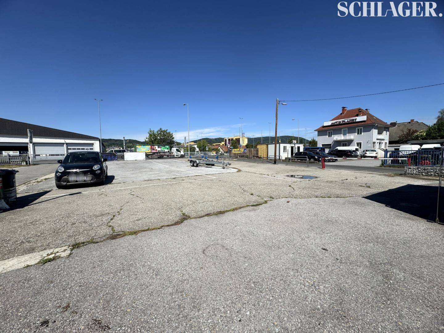 Großer asphaltierter Parkplatz mit Fahrzeugen und angrenzenden Gewerbegebäuden bei Sonnenschein.