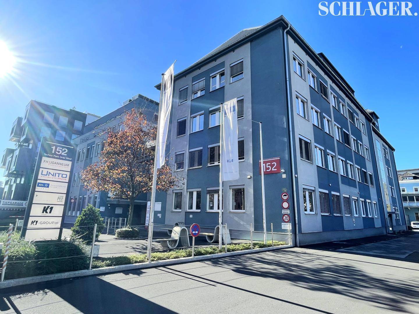 Modernes Bürogebäude mit blauer Fassade, großen Fenstern und Firmenflaggen unter strahlendem Himmel.