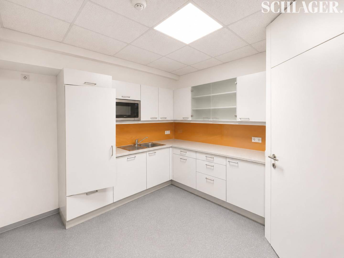 Moderne Einbauküche in Weiß mit Mikrowelle, Spüle und orangefarbener Rückwand in einem Büro.