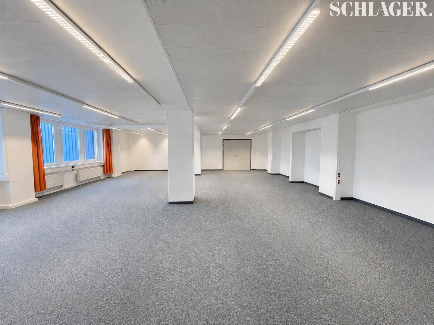 Langer, leerer Büroraum mit grauem Teppichboden, modernen Deckenleuchten und orangefarbenen Vorhängen.