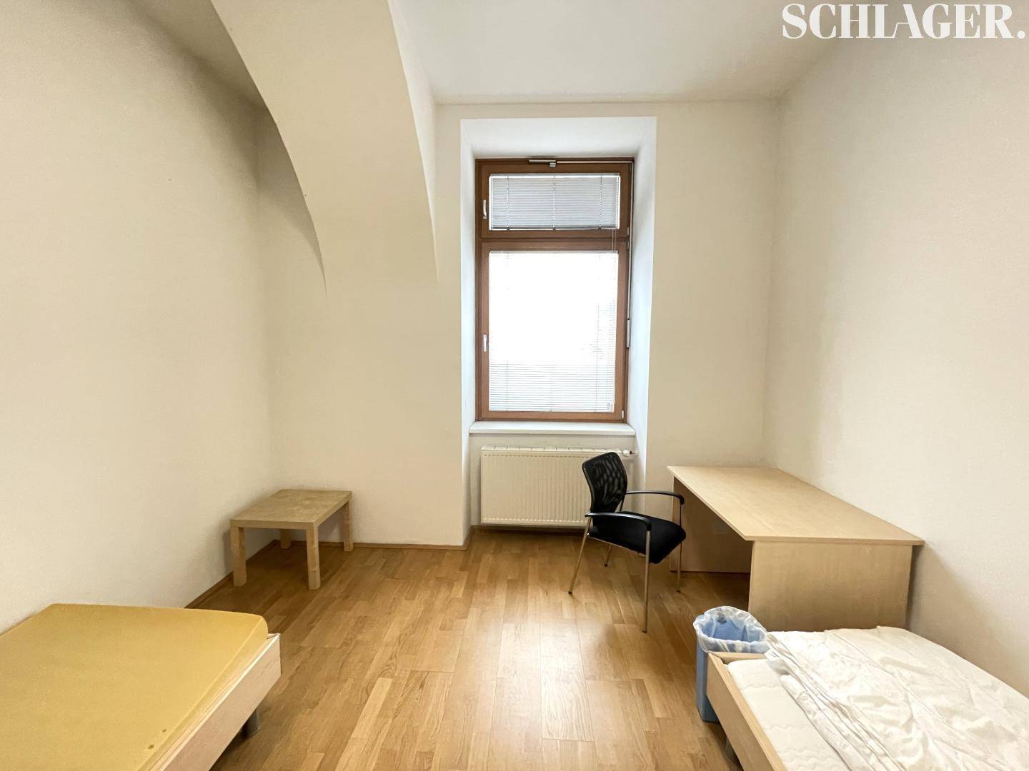 Einfaches Zimmer mit zwei Betten, Schreibtisch und einem Fenster mit Blick nach draußen.