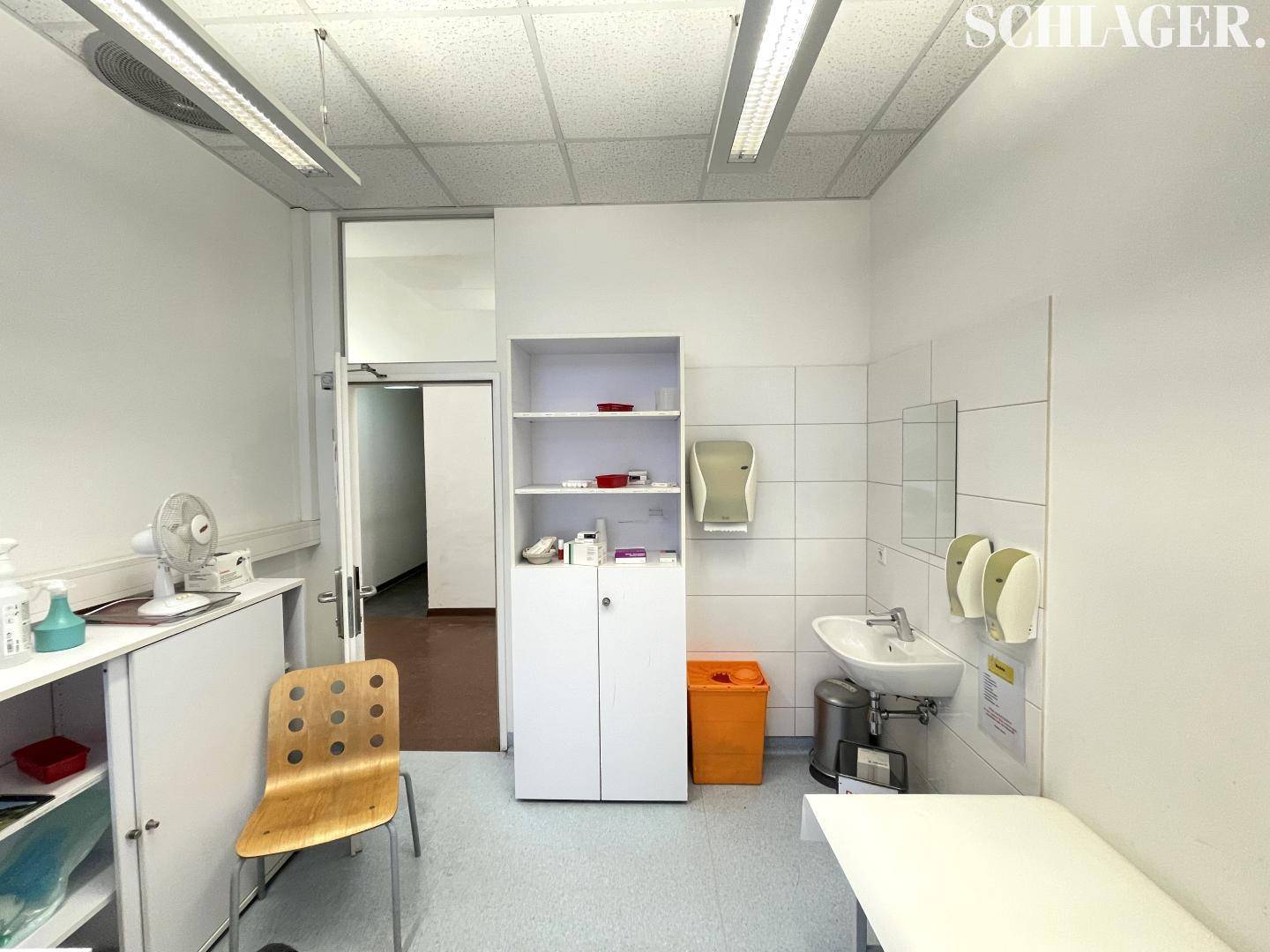 Helles Untersuchungszimmer mit Waschbecken, Schrank und Zugang zu einem weiteren Raum.