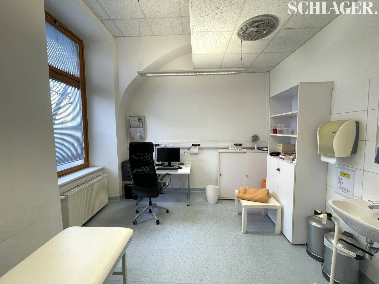 Untersuchungszimmer mit medizinischer Ausstattung, Schreibtisch und Fenster mit Jalousien.